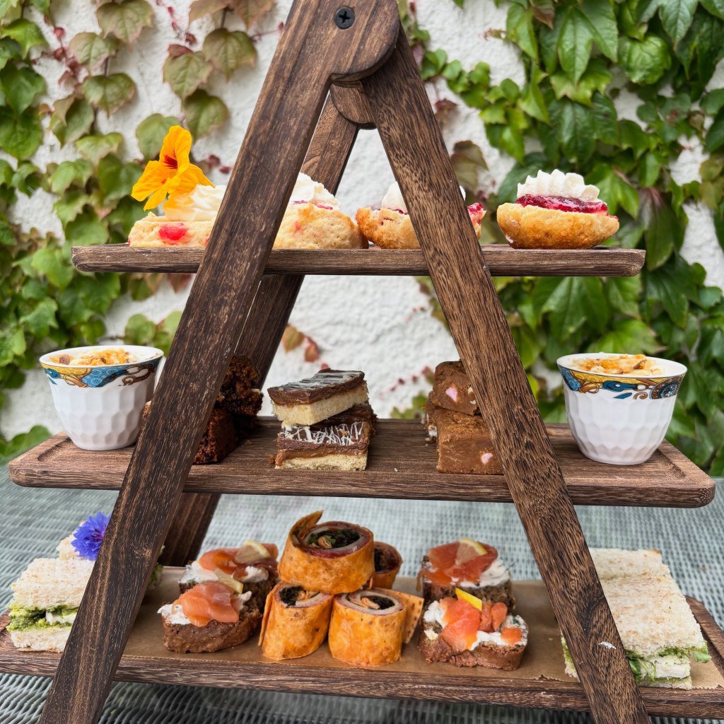 A multi-tiered wooden serving stand filled with a variety of pastries, desserts, and savory snacks, including cupcakes, brownies, and sandwiches, set against a backdrop of green foliage.