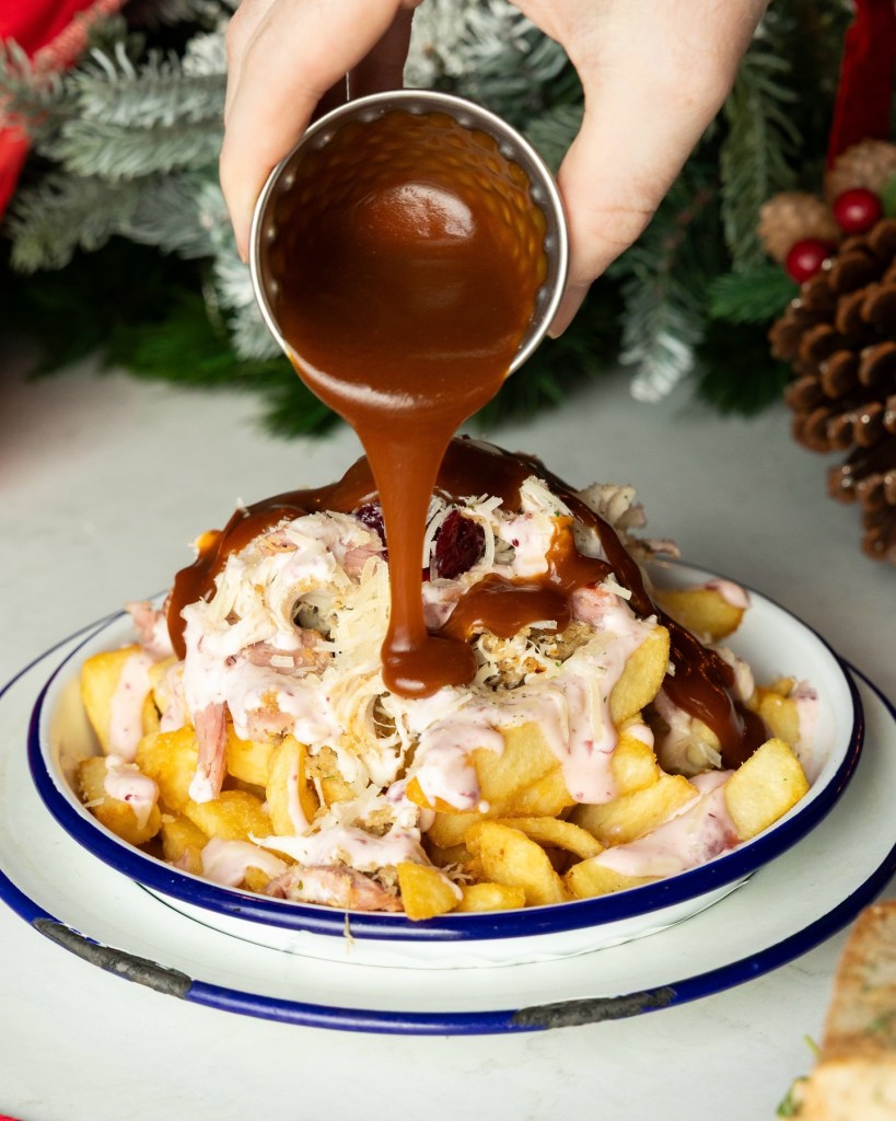 A close-up of a hand pouring gravy over a plate of festive fries topped with shredded turkey, cranberry sauce, and creamy toppings, set against a holiday-themed background.