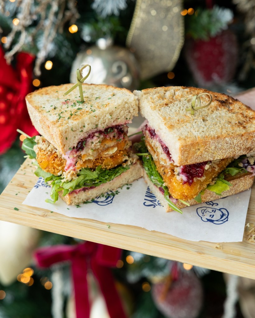 A festive sandwich with layers of lettuce, cranberry sauce, and a breaded filling, presented on a wooden board and set against a Christmas backdrop.