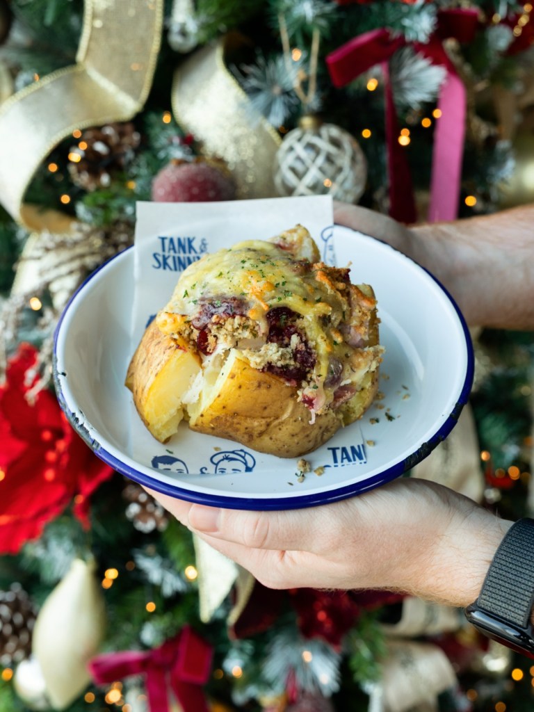 A hand holding a loaded baked potato with toppings, set against a festive Christmas backdrop featuring decorations and lights.