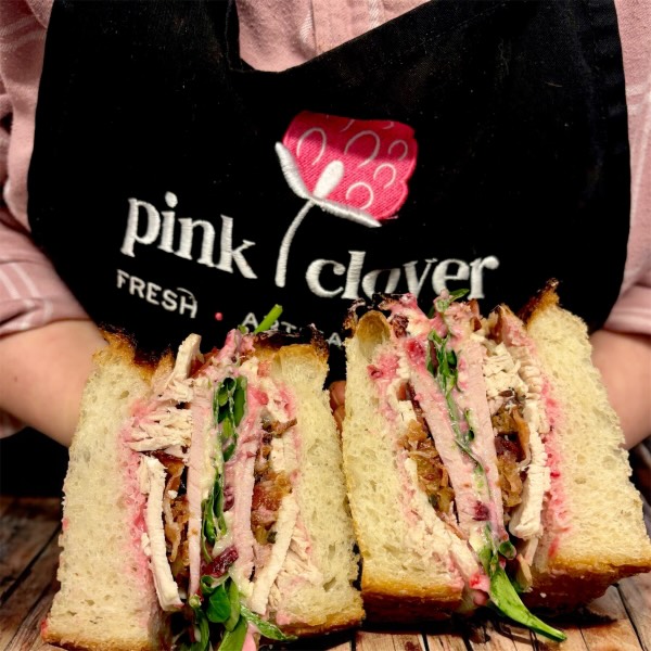 A close-up of a festive sandwich cut in half, showcasing layers of turkey, stuffing, and greens, with a person wearing a pink clover apron holding it.