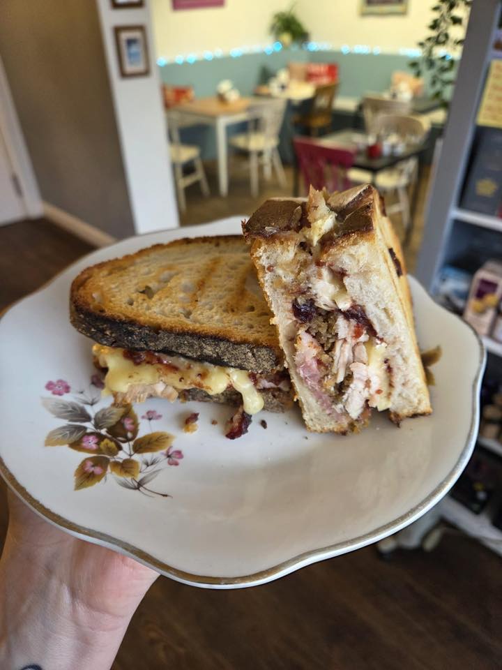 A close-up of a festive sandwich with layers of turkey, cheese, and cranberry sauce, served on a decorative plate in a cozy cafe setting.