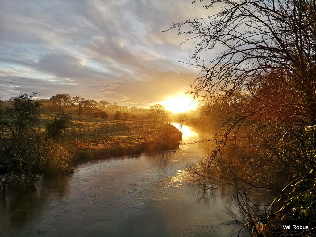 A serene winter landscape showing a river at sunrise, with mist rising from the water and trees lining the banks, creating a peaceful atmosphere.
