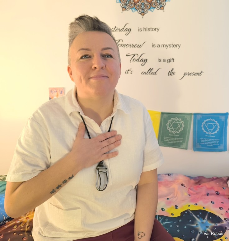 A person with short hair and a light shirt is smiling and placing their hand on their chest, in front of a wall with inspirational text and colorful decorative elements.
