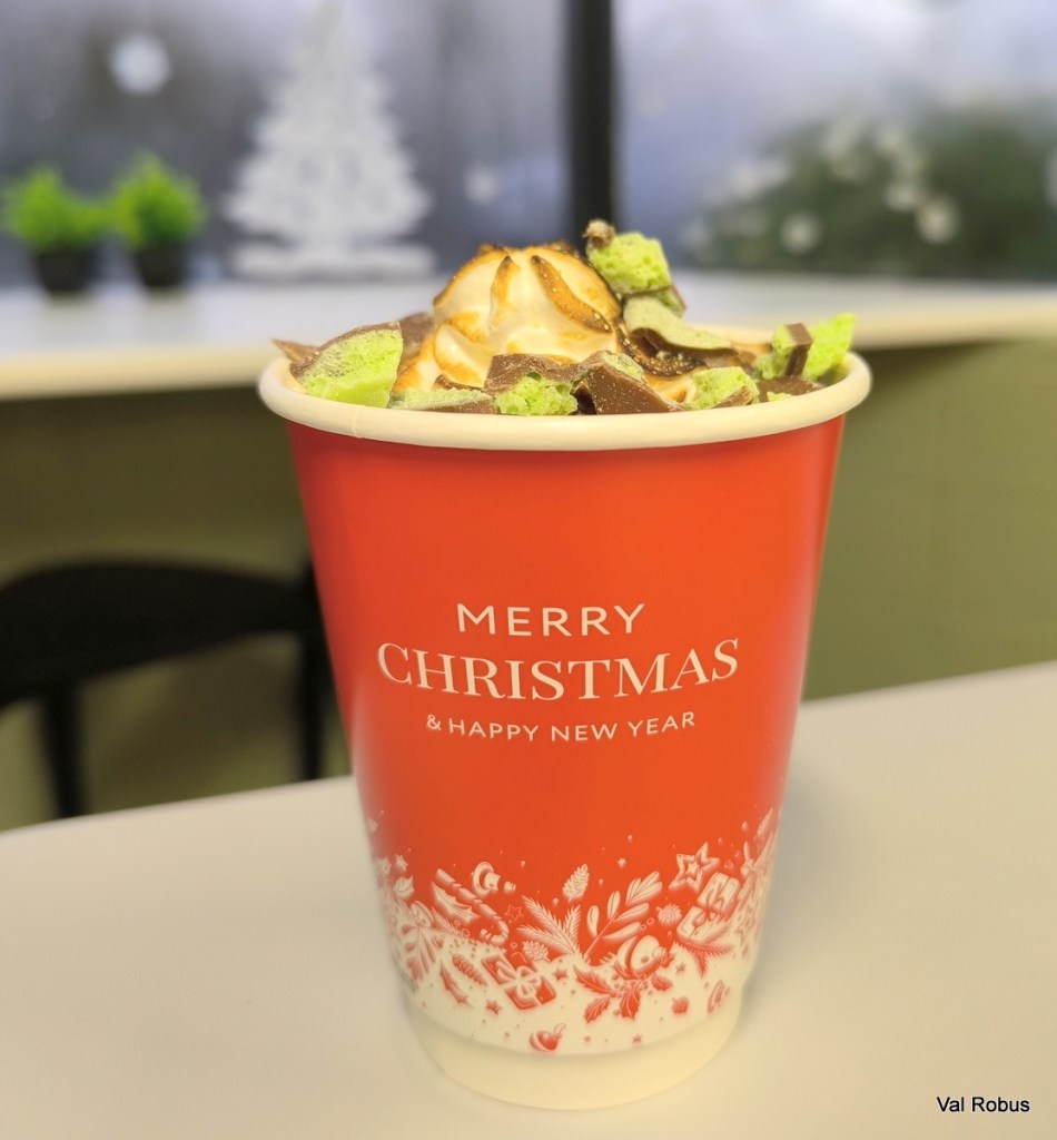 A festive red cup with 'MERRY CHRISTMAS & HAPPY NEW YEAR' printed on it, filled with a colorful dessert topped with whipped cream and chocolate pieces.