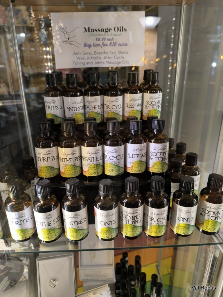 A display of massage oils in various scents, labeled with their benefits like Anti-Stress, Breathe Eze, and Sleep Well, arranged neatly on a glass shelf with a sign indicating prices and promotions.