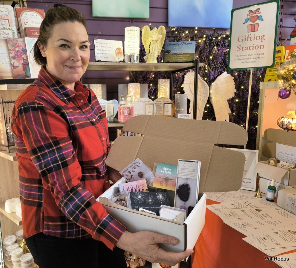 A woman in a red checkered shirt holds an open gift box filled with various items at a gifting station in a shop decorated for the holidays.