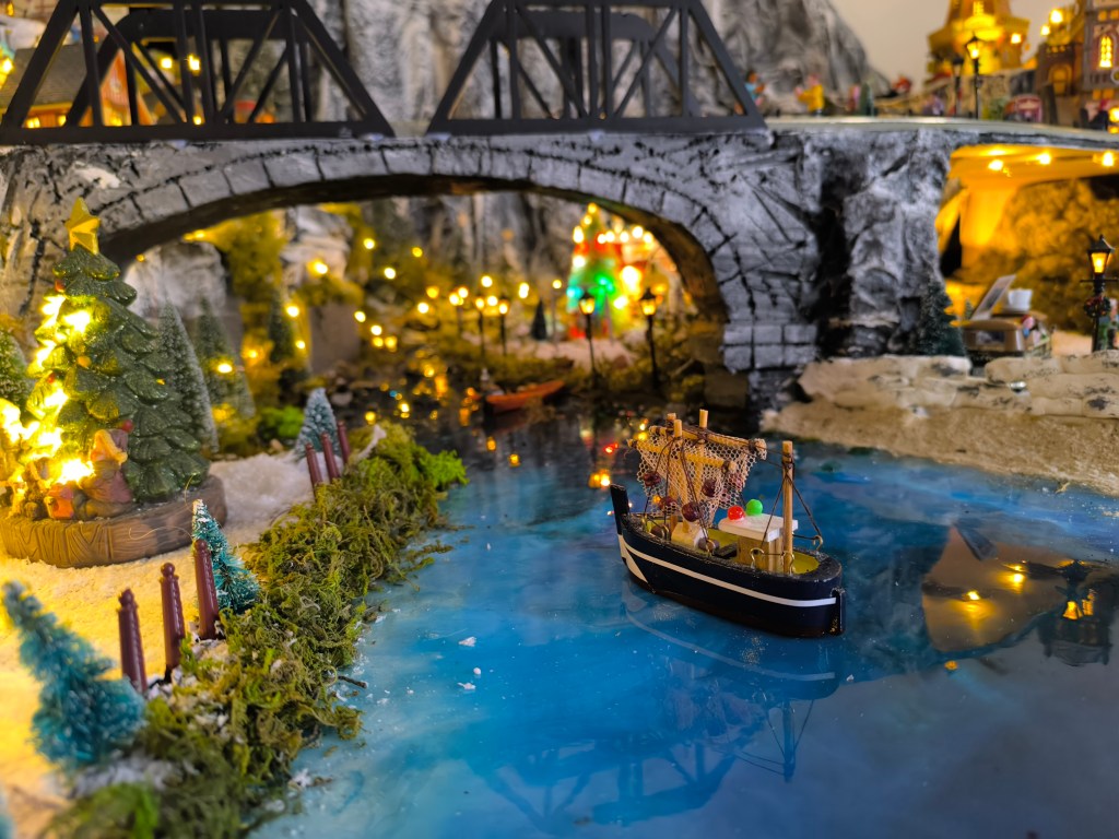 A miniature scene depicting a festive model village with a bridge, a decorated Christmas tree, and a small boat on a blue waterway.