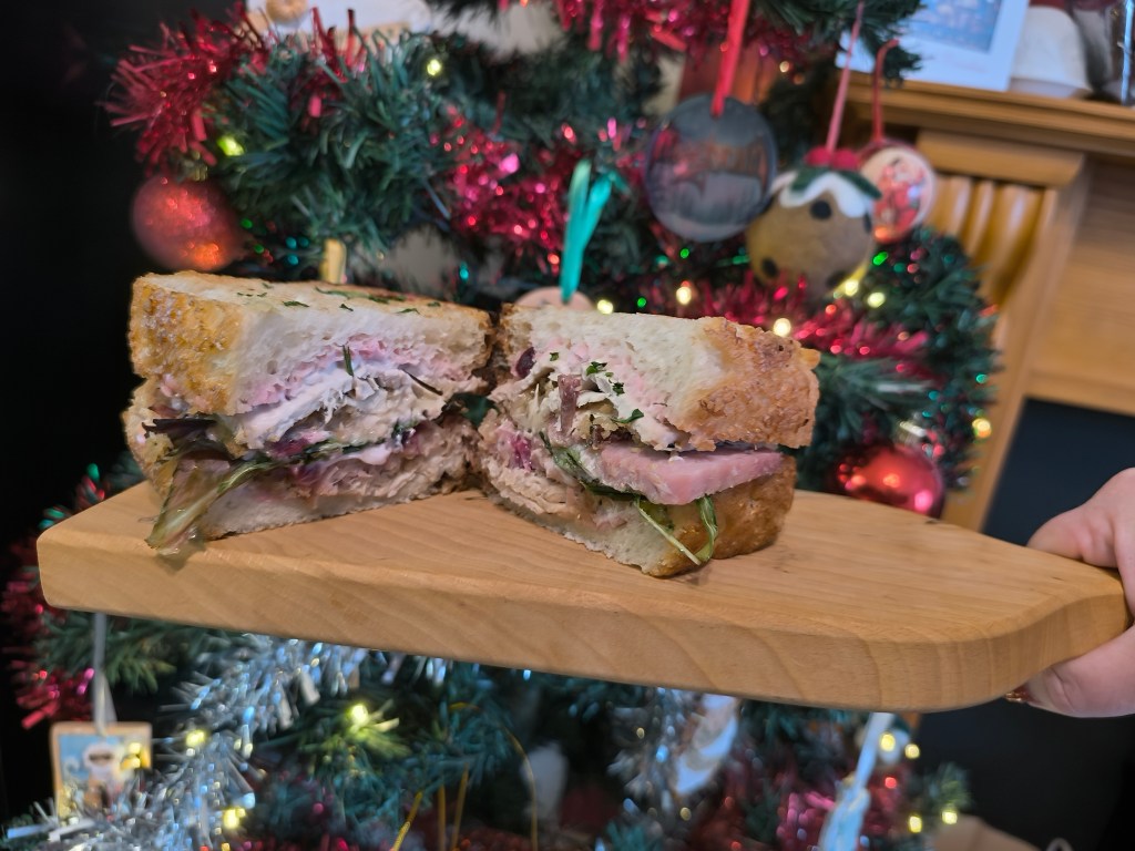 A hand holding a wooden platter with a sliced sandwich filled with meat and greens, in front of a festive Christmas tree adorned with colorful decorations.