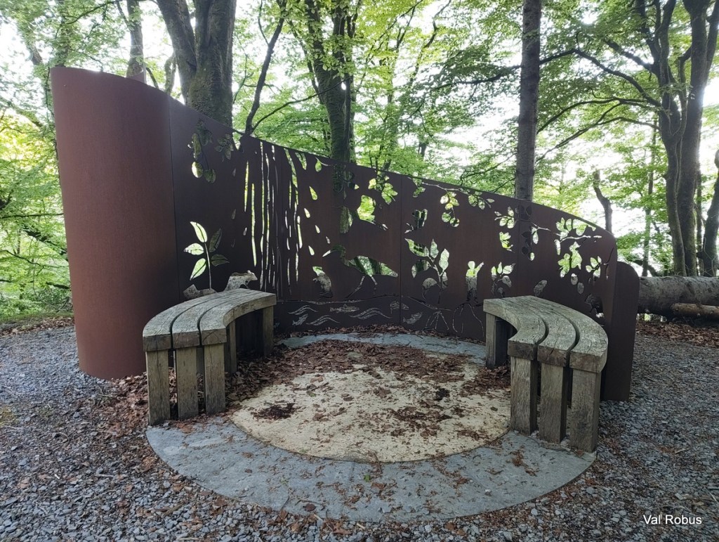 A unique outdoor seating area featuring wooden benches arranged in a circular shape, surrounded by a decorative metal screen with cut-out leaf and bird designs, set amidst a wooded environment.