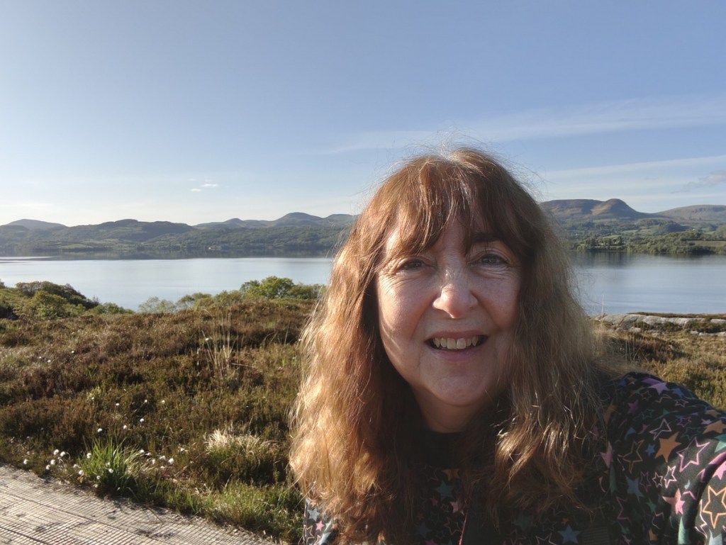 A smiling woman with long, wavy hair stands in front of a picturesque landscape featuring a calm lake and rolling hills under a clear blue sky.