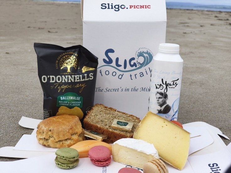 A picnic spread featuring a Sligo food trail box, a bag of O'Donnell's cheese crisps, slices of brown bread, cheese, a scone, colorful macarons, a banana, and a bottle of water with a portrait of W.B. Yeats, set against a sandy beach.