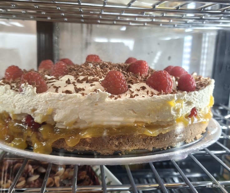 A layered cake featuring a creamy white topping, garnished with fresh raspberries and chocolate shavings, displayed in a refrigerated case.