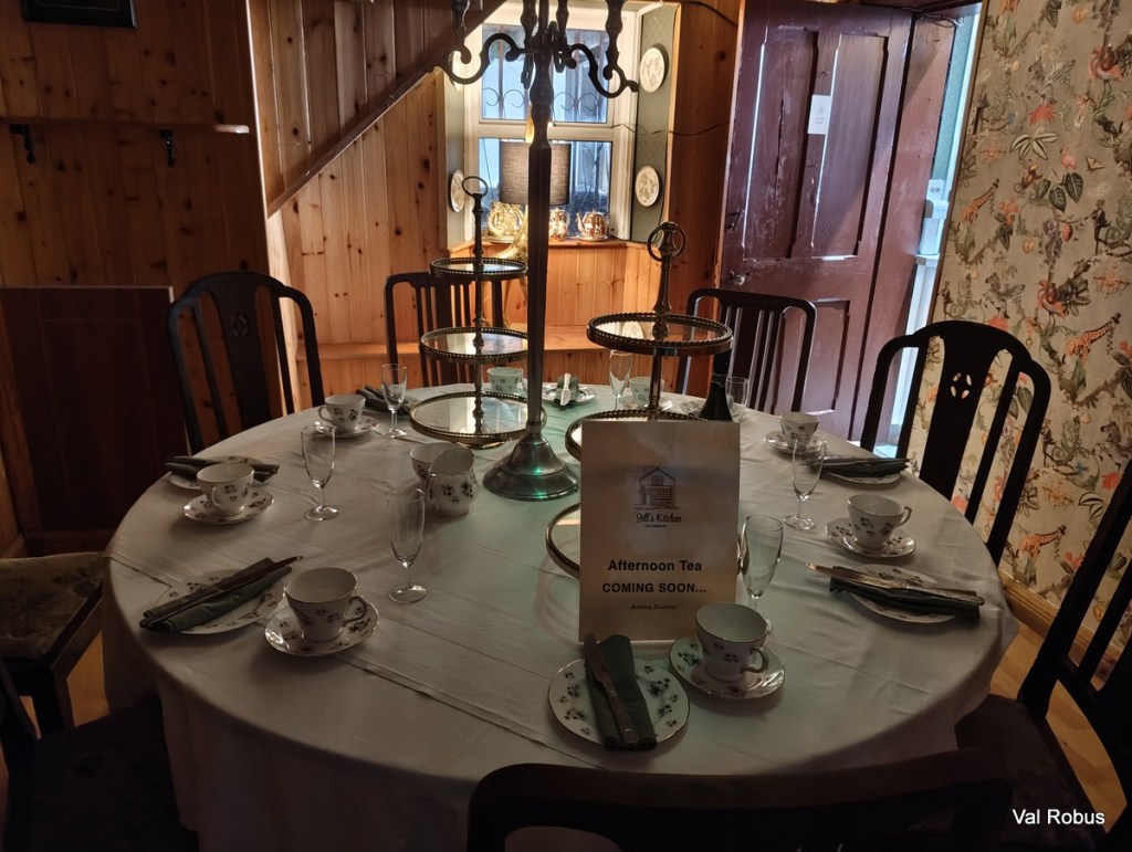 A circular dining table set for afternoon tea, featuring floral-patterned china cups and plates, glassware, and a three-tiered serving stand. A sign reads 'Afternoon Tea COMING SOON...'. The background includes wooden walls and a partially open door.