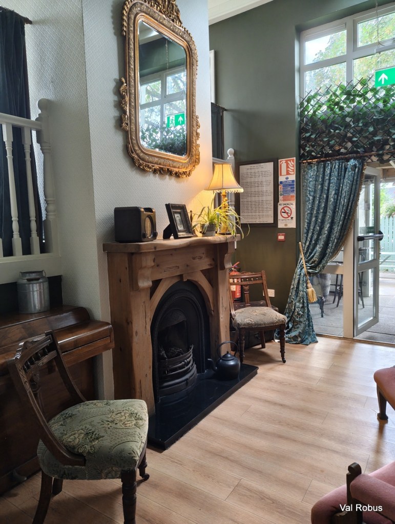 Interior of a cozy room featuring a vintage mantelpiece, a large ornate mirror, a sofa, and wooden chairs. There is a plant and a lamp on the mantel, along with a small radio and a framed photo, creating a warm and inviting atmosphere.