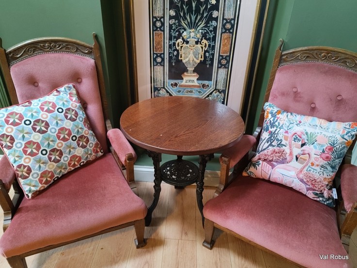 A cozy seating area featuring two vintage pink upholstered armchairs with decorative pillows, positioned around a small round wooden table. A colorful wall art piece is visible behind the chairs, set against a green wall.