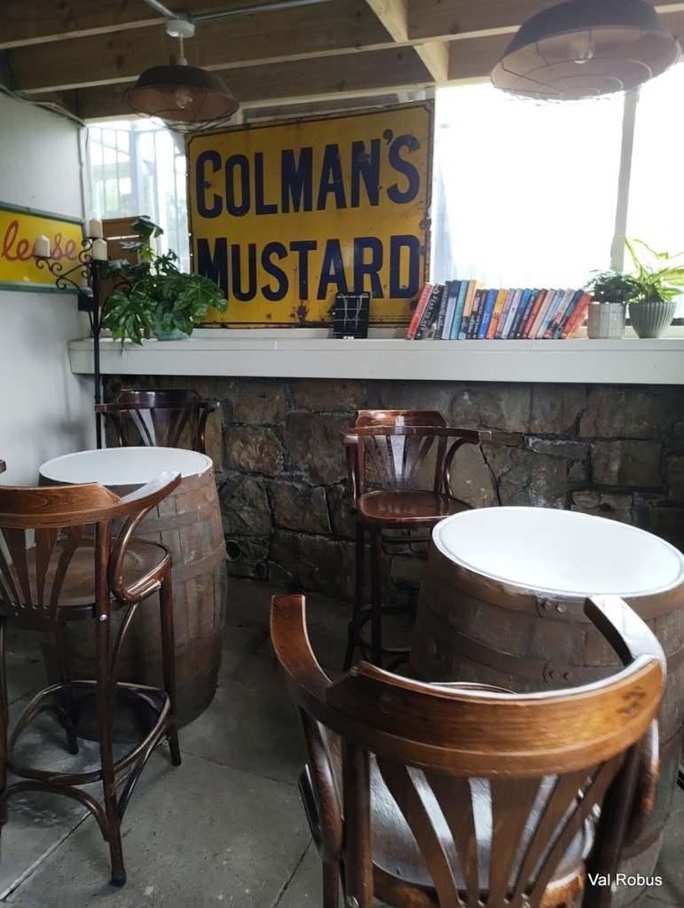 Interior of a rustic café featuring wooden chairs and tables, with an old sign reading 'COLMAN'S MUSTARD' on the wall, surrounded by books and plants.