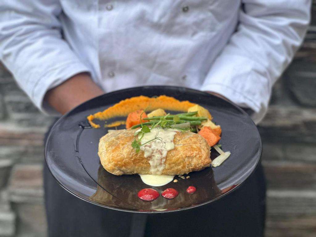 A chef holding a black plate featuring a pastry wrapped dish with a creamy sauce, surrounded by colorful vegetables including carrots and asparagus.