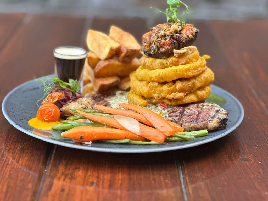 A gourmet dish featuring a grilled steak, crispy onion rings, fried potatoes, cooked carrots, and green beans, served with a small cup of dark sauce on a black plate.