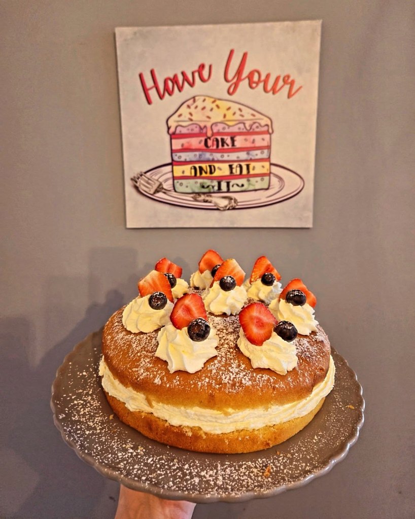 A freshly baked cake decorated with whipped cream, strawberries, and blueberries, served on a silver plate, with a whimsical cake-themed sign in the background that says 'Have Your Cake And Eat It.'
