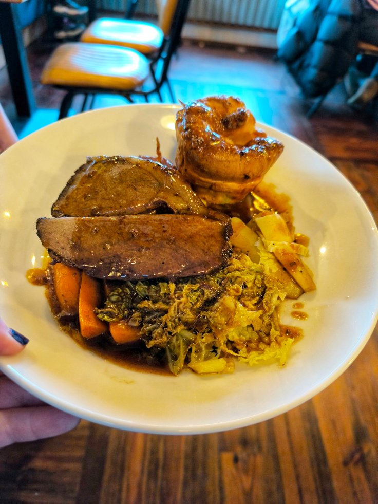 A plate of sliced cooked meat, accompanied by vegetables including carrots and cabbage, and a roasted pastry on top, held in a person's hand.