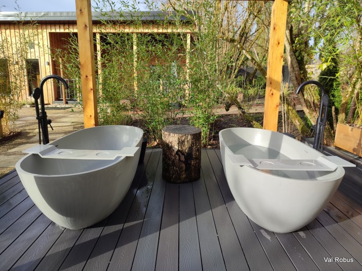 Two modern soaking tubs placed on a wooden deck surrounded by greenery, with a wooden stump in between them.