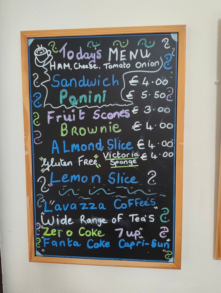 A chalkboard menu listing food and drink items including Sandwich, Panini, Fruit Scones, Brownie, Almond Slice, Lemon Slice, Lavazza Coffee, a range of teas, and soft drinks like Zero Coke and Fanta.