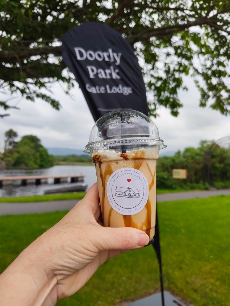 A person holding a caramel-drizzled iced coffee drink with a logo on the cup, in front of a black flag displaying 'Doorly Park Gate Lodge' and a scenic landscape in the background.