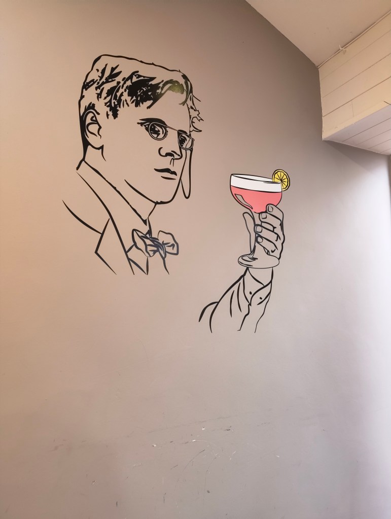 A wall mural depicting a stylized man with glasses and a bow tie, holding a pink cocktail with a lemon slice.