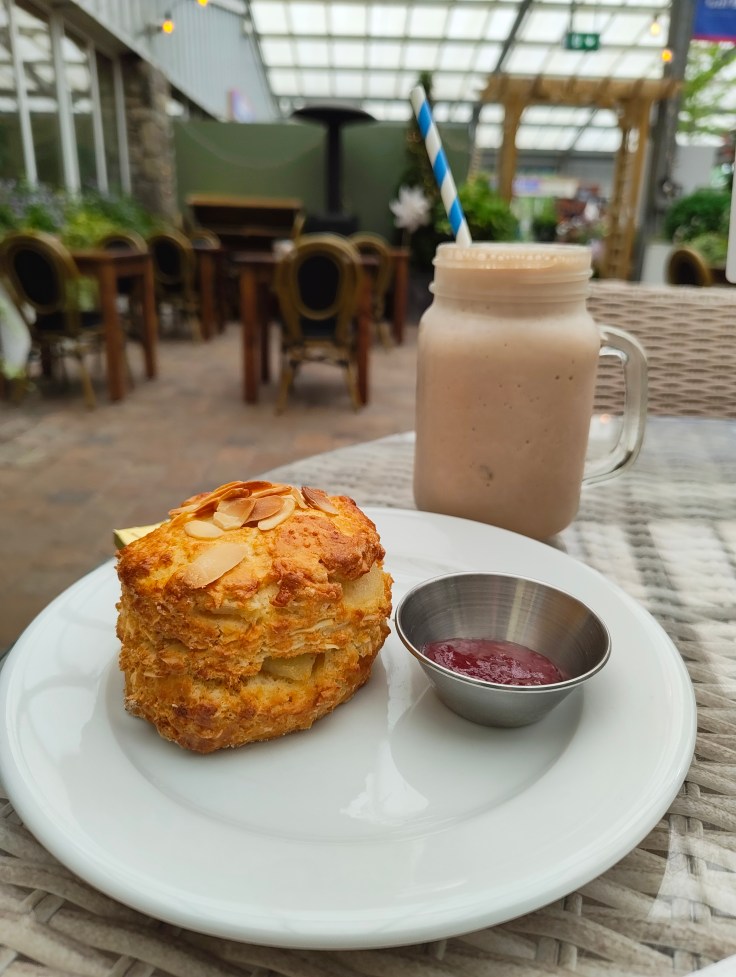 A freshly baked scone topped with sliced almonds, served on a white plate alongside a small dish of jam and a jar of smoothie with a blue and white striped straw.