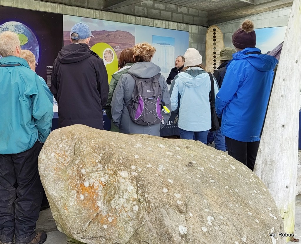 A group of people listening to a speaker in an indoor setting, with a large rock in the foreground and informative panels on the wall.