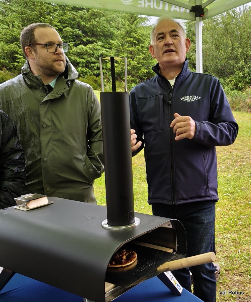 Two men standing under a tent, one explaining while the other listens. They are near a portable pizza oven displaying a freshly made pizza.
