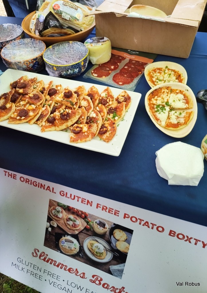 A table display featuring gluten-free potato boxty, topped with various ingredients including salami and cheese, along with packaging for additional food items and a promotional sign for 'Slimmers Boxty' highlighting gluten-free, low-fat, and vegan options.