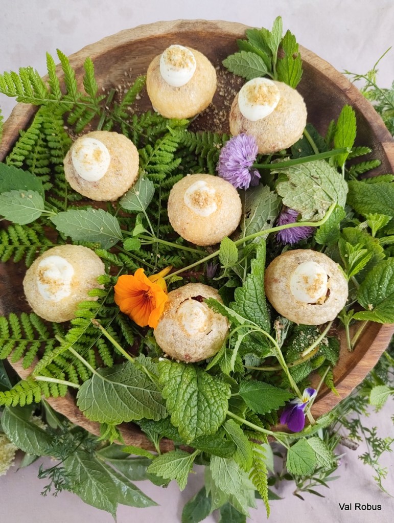 A wooden bowl filled with round, baked treats topped with white icing, surrounded by fresh greenery and colorful edible flowers.