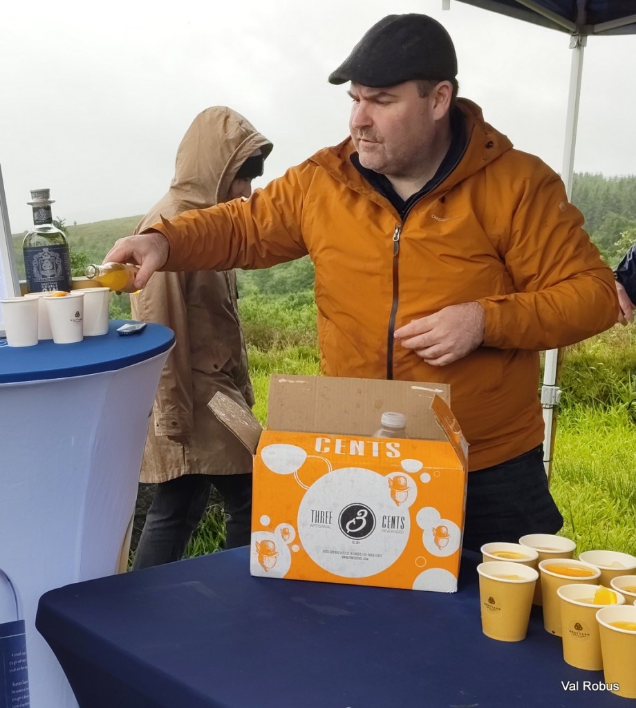 A man in an orange jacket pours a drink from a bottle into a paper cup at an outdoor event, with a box labeled 'Three Cents' nearby.