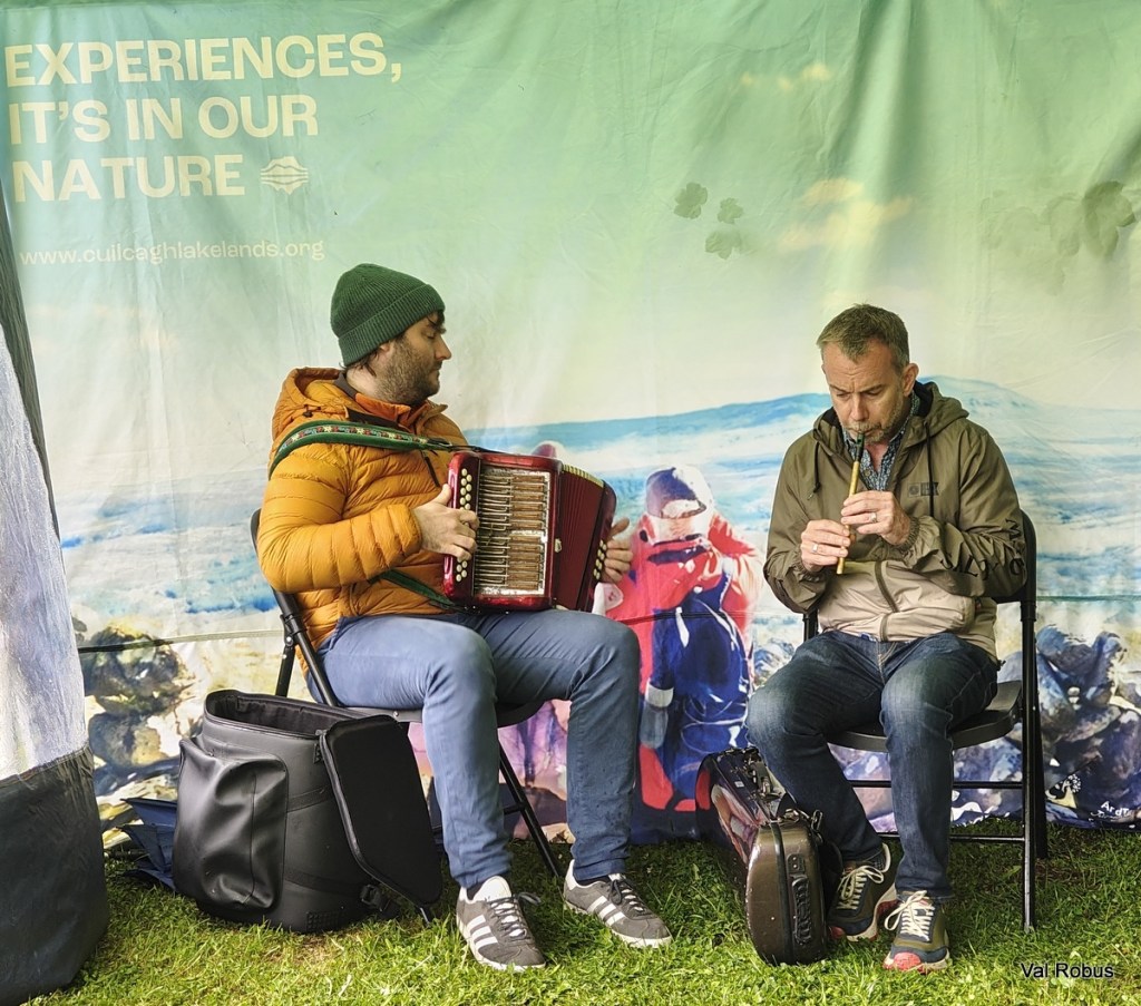 Two musicians seated in front of a colorful backdrop, one playing an accordion and the other playing a flute.