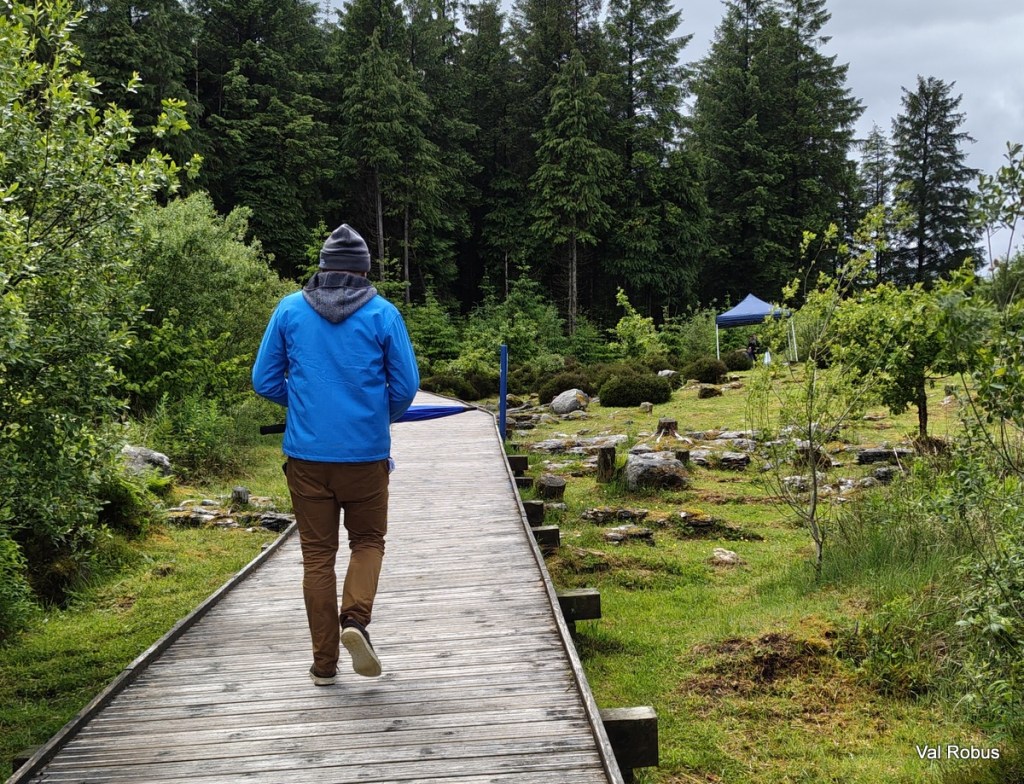 A person walking along a wooden pathway in a forested area, wearing a blue jacket and a beanie. Surrounding greenery includes trees, shrubs, and rocks.