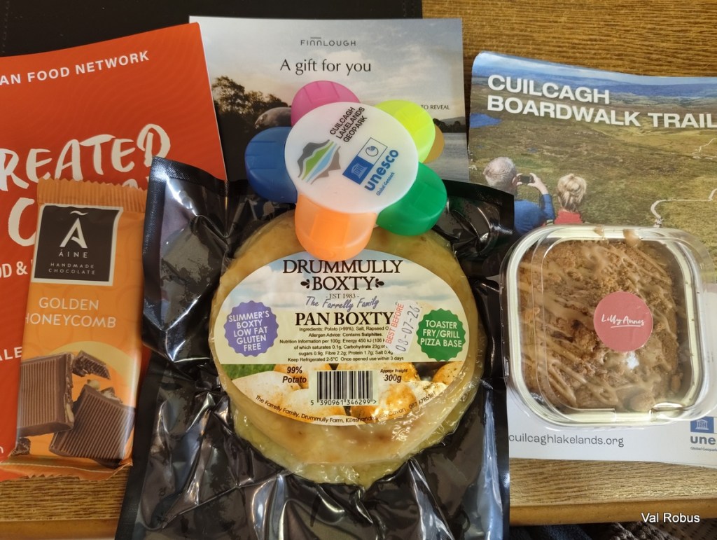 A selection of local food items including a bar of golden honeycomb chocolate, a gluten-free potato boxty labeled as 'Drummully Boxty', a small dessert from 'Lily Anne's', and brochures related to the Cuilcagh Boardwalk Trail and a gift program.