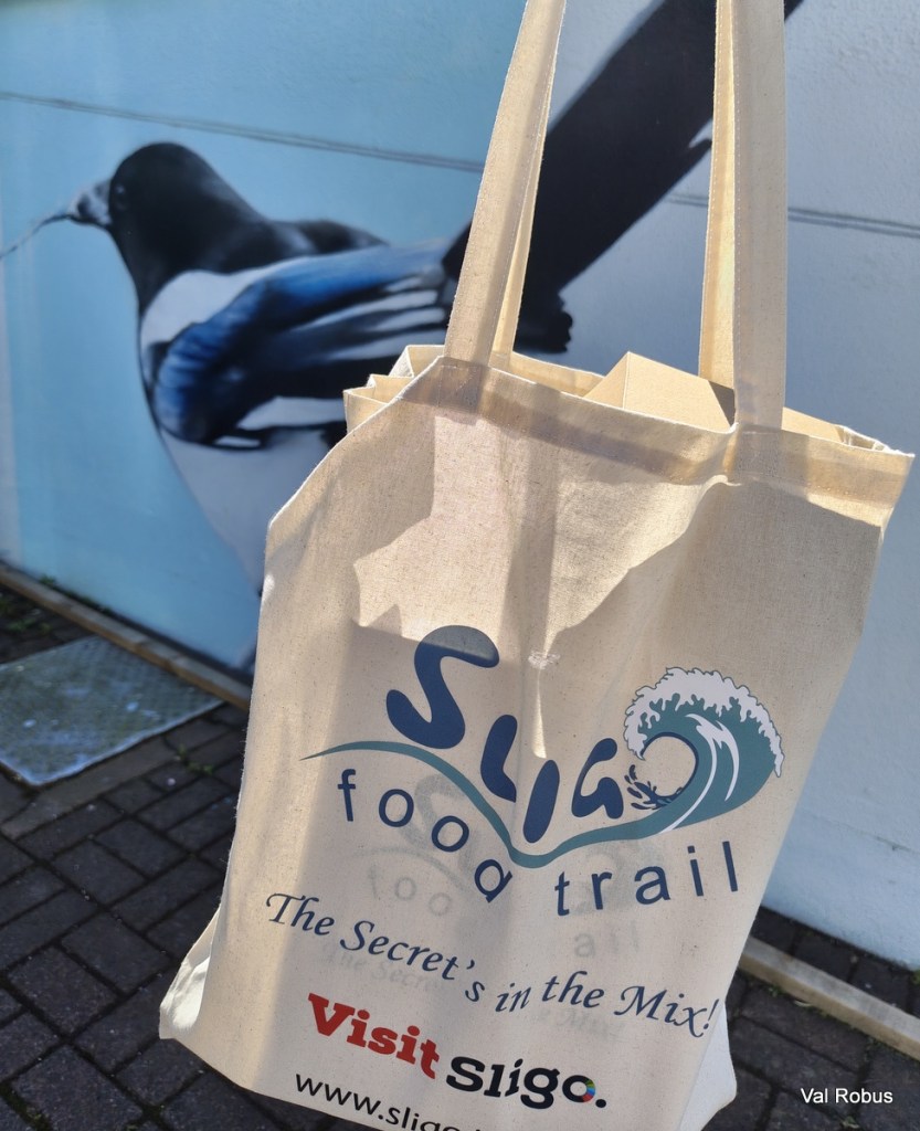 A beige tote bag featuring the text 'Sligo food trail' and a wave design, with a background mural of a bird.