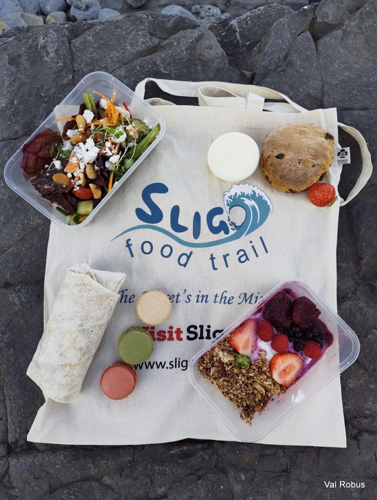 A picnic setup featuring various food items on rocks, including a salad in a plastic container, a wrapped tortilla, a container of yogurt with granola and berries, and colorful macarons. A reusable tote bag with 'Sligo Food Trail' branding is present.