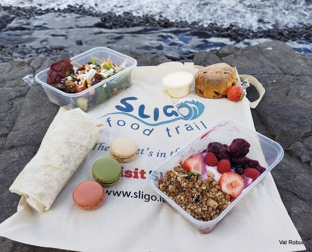A picnic spread featuring a salad in a container, a wrap, a scone, assorted macarons in colors, a yogurt parfait with berries, and a small container of sauce, all arranged on a Sligo food trail tote bag by the seaside.