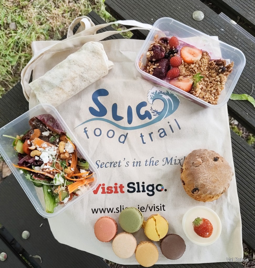 A tote bag featuring 'Sligo Food Trail' lays on wooden planks, displaying a variety of food items. A wrapped burrito is visible alongside a container of colorful salad with nuts and dressing, a cup of yogurt topped with berries and granola, a scone, and an array of colorful macarons.