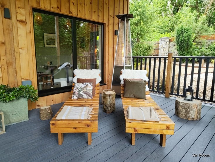 Two wooden lounging chairs with decorative pillows on a porch, accompanied by a heater and a small table, surrounded by greenery and a wooden structure.