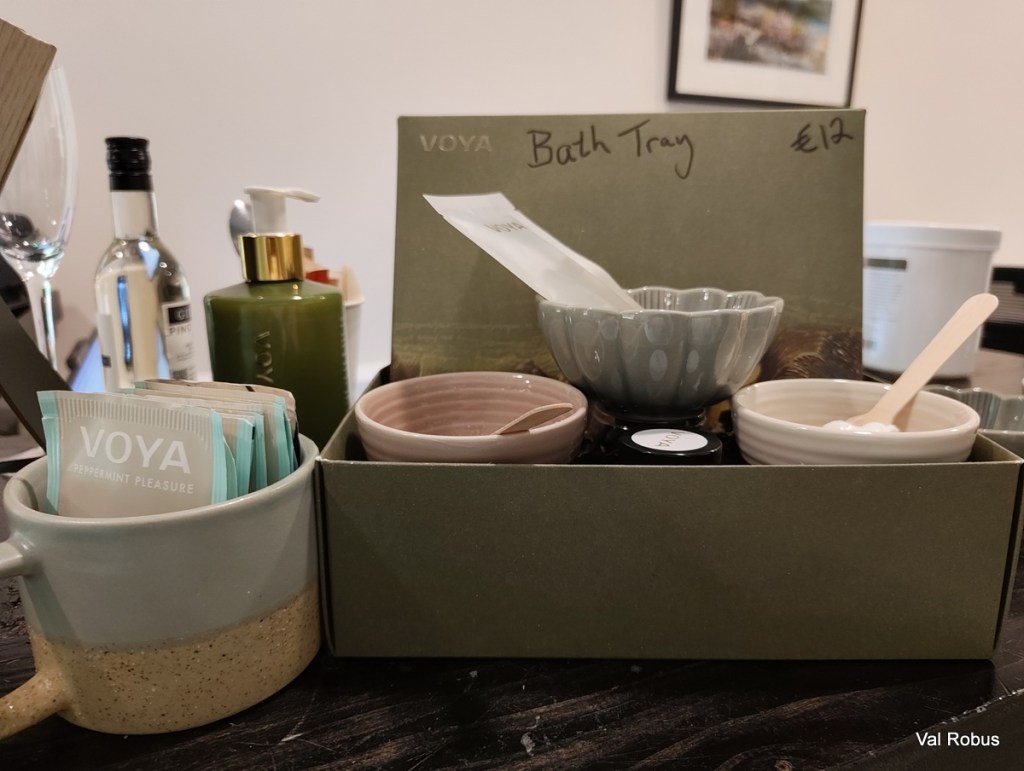 A decorative bath tray containing various skincare products, including a green bottle and small bowls. There are also packets labeled 'VOYA Peppermint Pleasure' and a small wooden spoon.