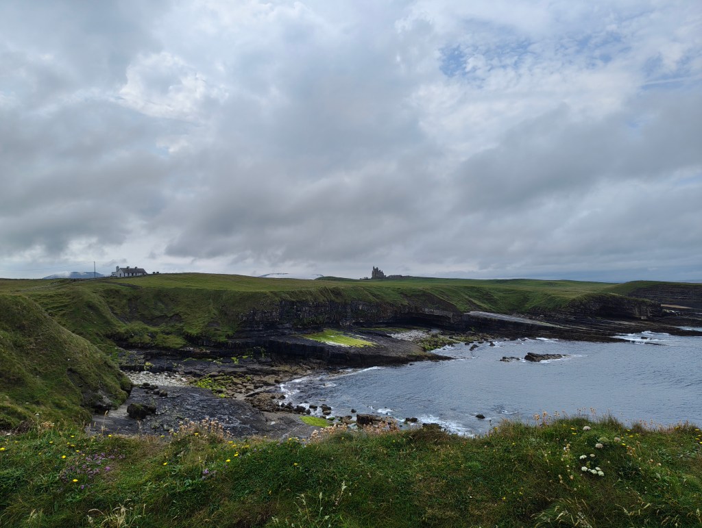 A scenic coastal view featuring green grassy hills, rocky shores, and a calm ocean under a cloudy sky.