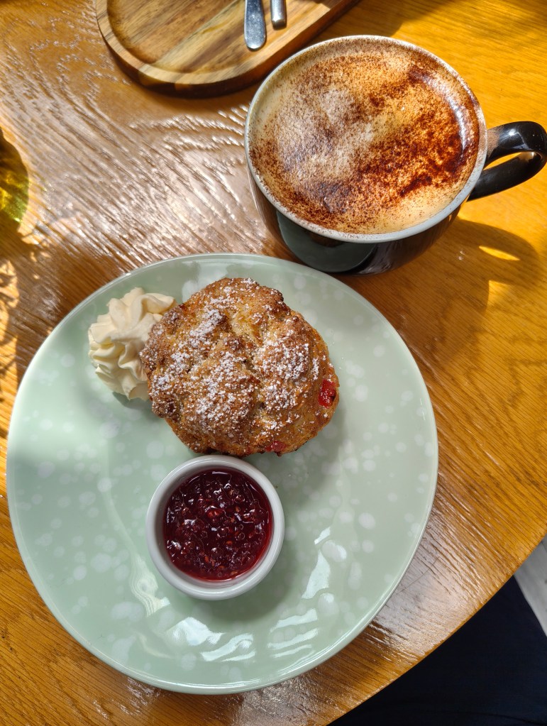 A plate with a muffin topped with powdered sugar, a dollop of whipped cream, and a small bowl of red jam, accompanied by a cup of cappuccino on a wooden table.