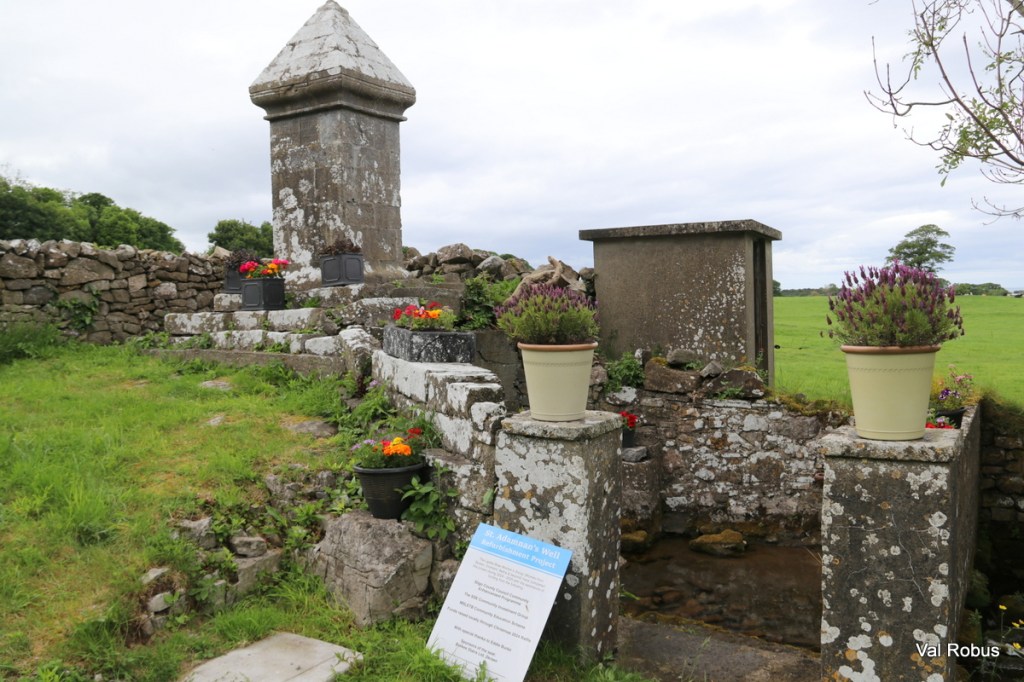 A historic well monument surrounded by stone walls, decorated with flower pots. In the foreground, a sign provides information about the site.