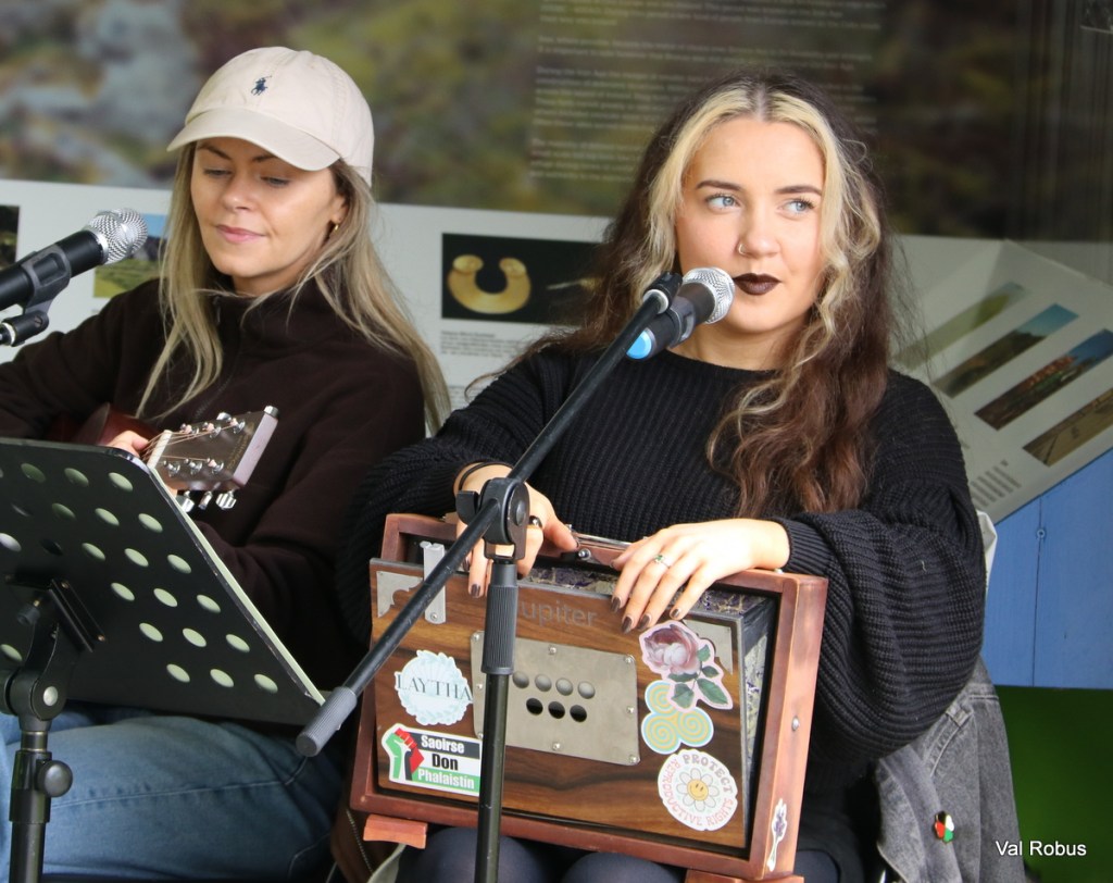 Two women performing music together, one playing guitar and the other playing a small musical instrument while singing. They are seated with a microphone and decorative stickers on the instruments.