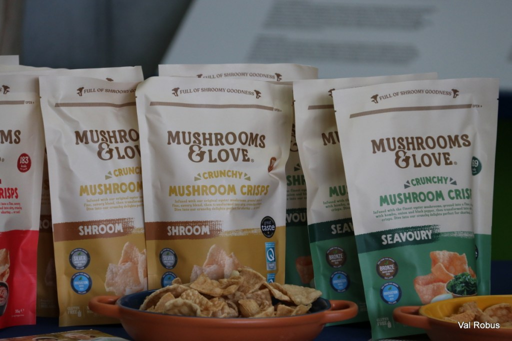 An assortment of mushroom crisp bags from the brand Mushrooms & Love, featuring flavors like 'Shroom' and 'Seavory', displayed on a table next to a bowl of mushroom snacks.
