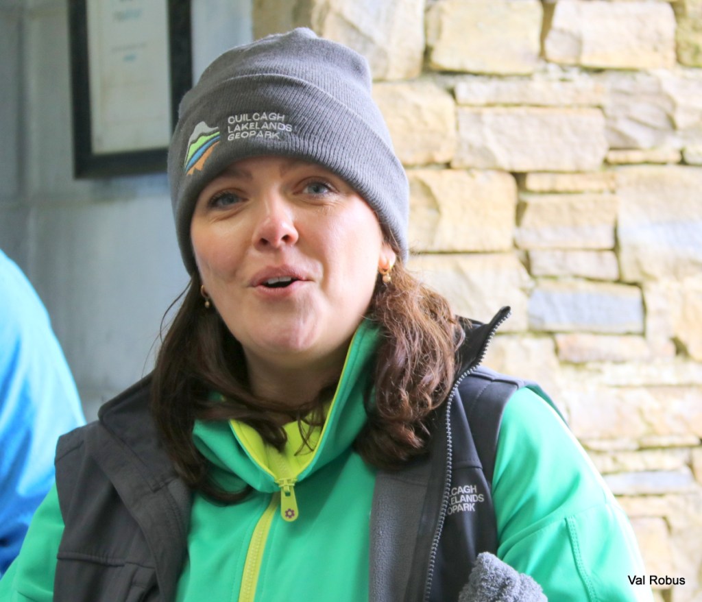 A woman with long brown hair is talking, wearing a gray beanie and a bright green jacket. She appears engaged and animated, with a stone wall in the background.